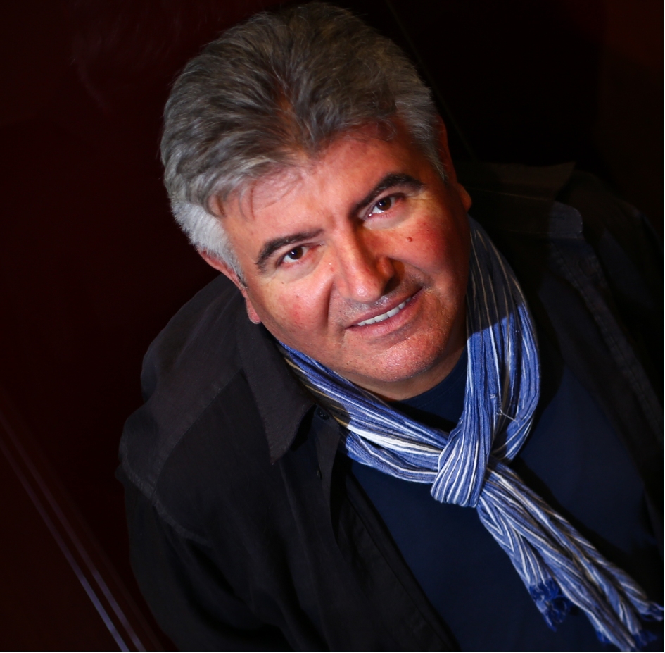 Ballet dancer in black leotard and white tulle skirt raises arms overhead, photographed from behind against textured white wall.