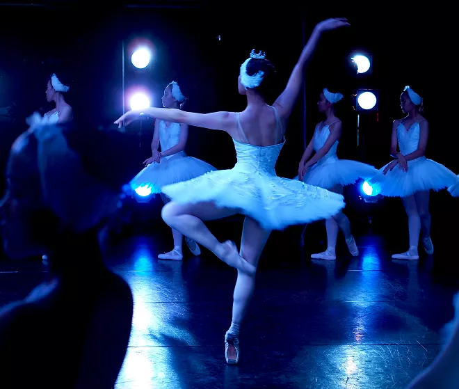 Ballet dancer in white tutu performs en pointe center stage with ensemble in blue lighting and stage lights.