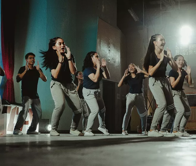 Ballet dancer in white tutu performs en pointe center stage with ensemble in blue lighting and stage lights.