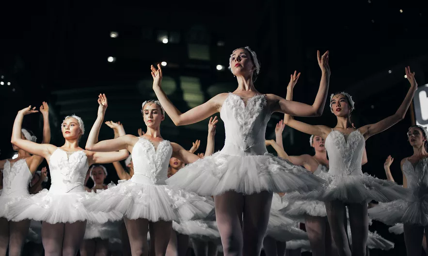 Ballet dancers in white tutus performing with raised arms on a dark stage with audience visible
