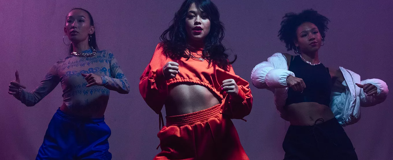 Three dancers in colorful crop tops perform contemporary choreography against a purple-lit studio backdrop.