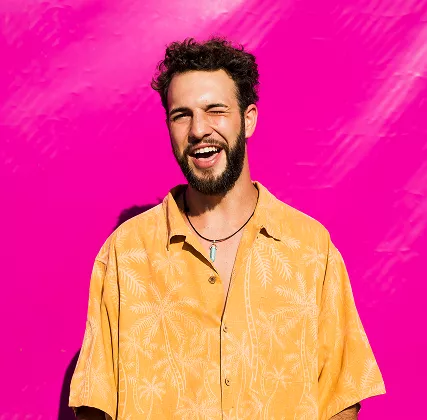 Bearded man in yellow floral shirt laughs joyfully against vibrant hot pink studio background.