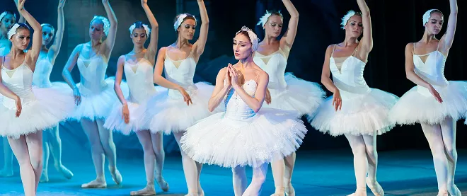 Ballet corps in white tutus performs synchronized movements with lead dancer centered, against turquoise-lit stage backdrop.