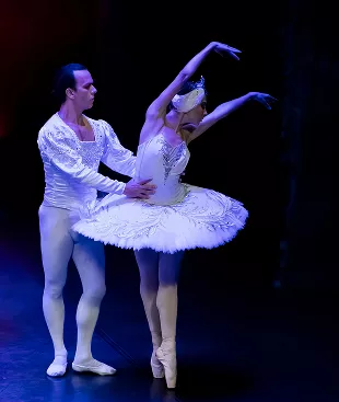 Male and female ballet dancers perform pas de deux in white costumes under purple stage lighting, ballerina en pointe.