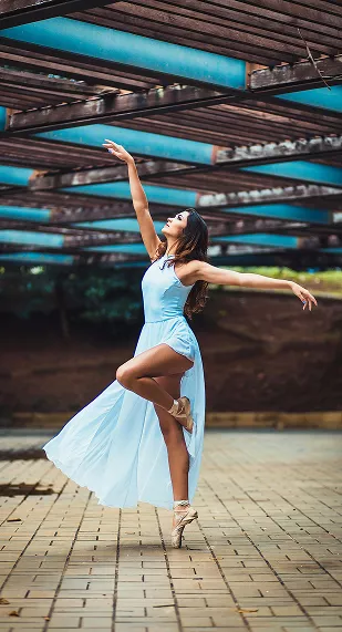 Ballet dancer in flowing light blue dress strikes elegant pose outdoors on brick pavement beneath modern architectural beams.