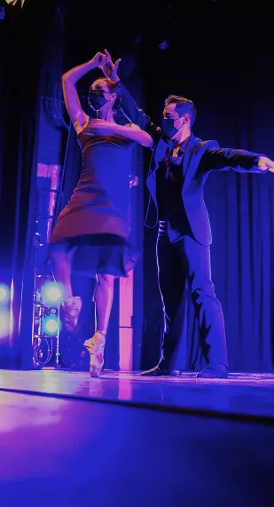 Ballet partners perform on stage under purple lighting, ballerina en pointe with arms raised, male dancer supporting beside her.