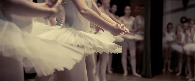 Ballet dancers in white tutus perform synchronized arabesque movements on stage, with other dancers visible observing in background.