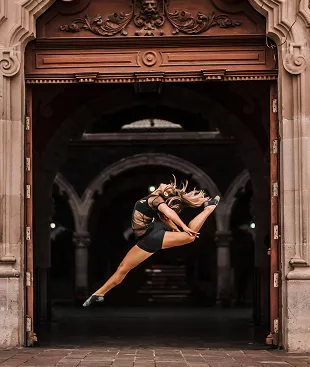 Dancer performs dramatic leap in black outfit framed by ornate architectural doorway with classical carved details and arches